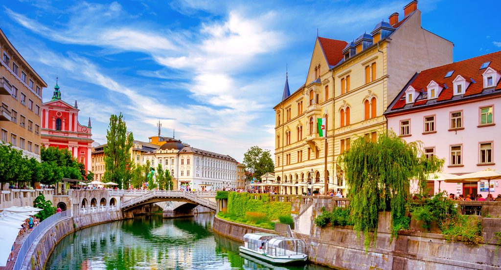 Charming riverside buildings with red rooftops and a canal boat gliding under a stone bridge in Ljubljana’s Old Town.