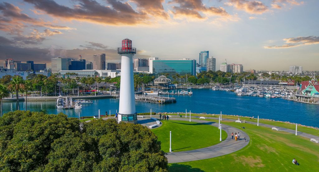 Scenic Long Beach waterfront with a white lighthouse, marina, and downtown skyline bathed in warm evening light.