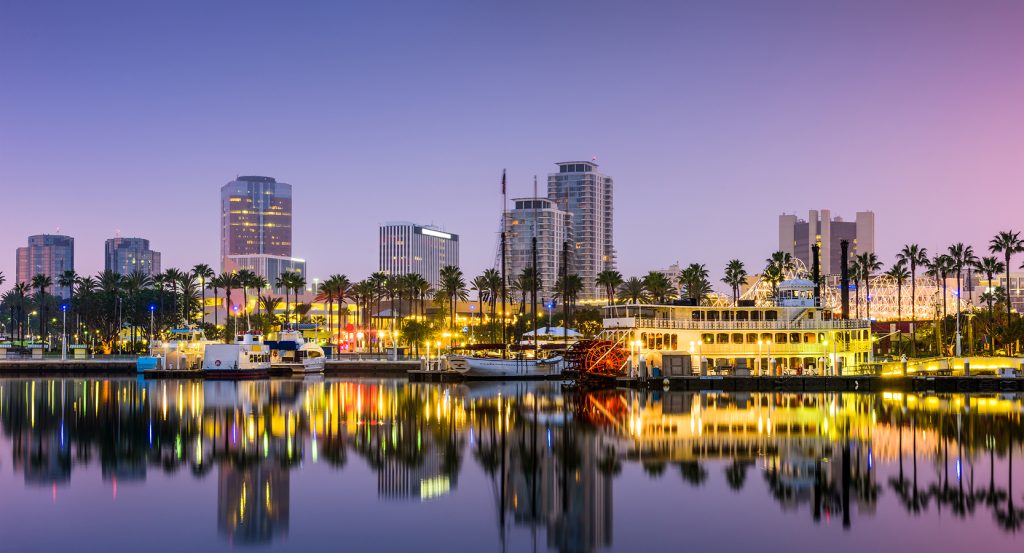 Long Beach’s skyline glows at twilight, mirrored in the calm harbor waters, with palm trees and docked yachts setting the SoCal coastal tone.