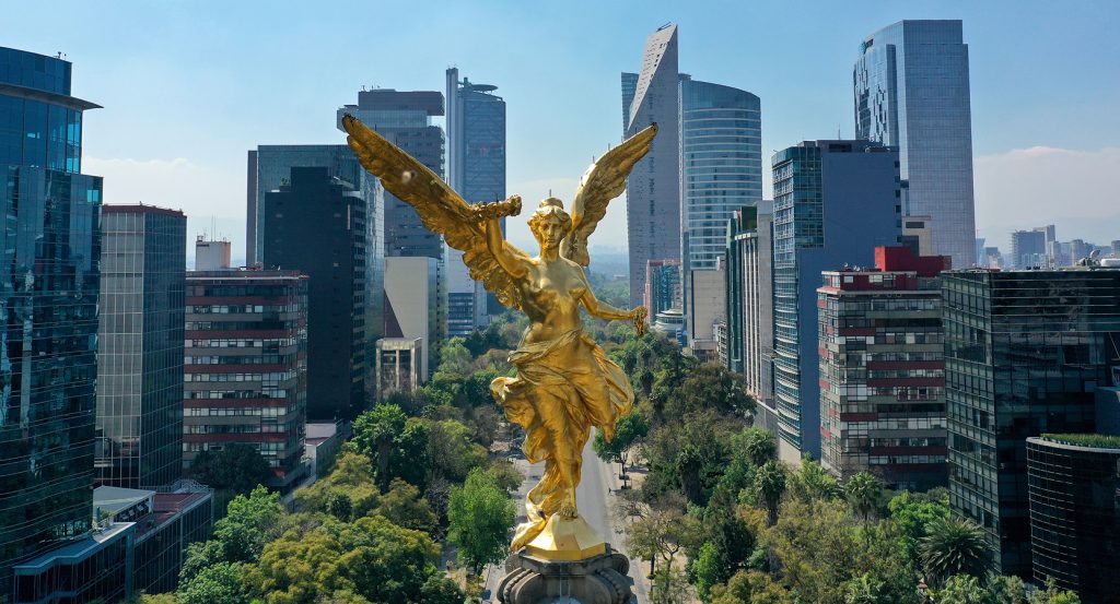 La estatua dorada del Ángel de la Independencia en la Ciudad de México, rodeada de rascacielos modernos y avenidas arboladas.