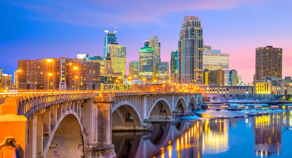 Twilight view of the Minneapolis skyline with a glowing bridge stretching over the calm Mississippi River, reflecting the city lights in the water.