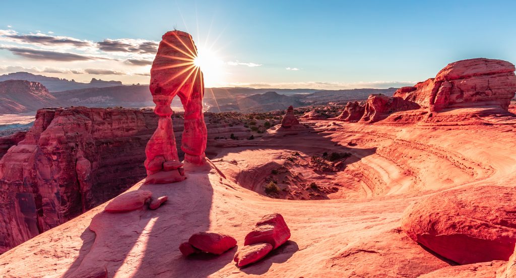 Sun peeking through Delicate Arch at Arches National Park in Moab, Utah, casting long shadows across red sandstone formations at golden hour.