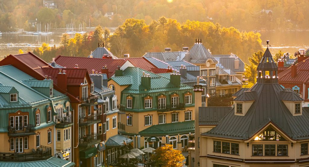 Charming alpine-style buildings with colorful rooftops in Mont-Tremblant Village, nestled by a lake and surrounded by glowing autumn foliage.