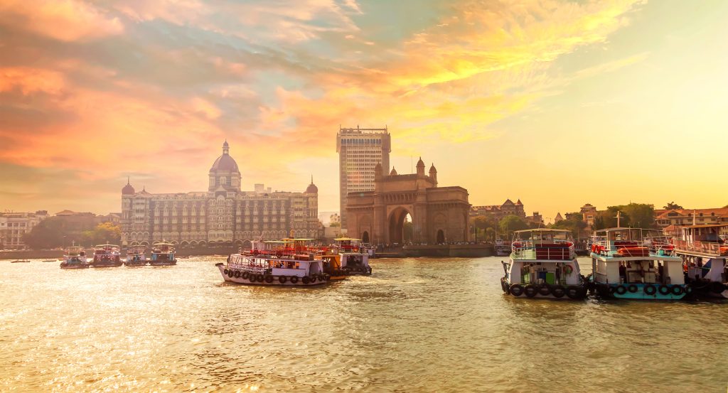 View of the Gateway of India at sunrise with ferry boats docked in the Arabian Sea and the iconic Taj Mahal Palace Hotel in the background.