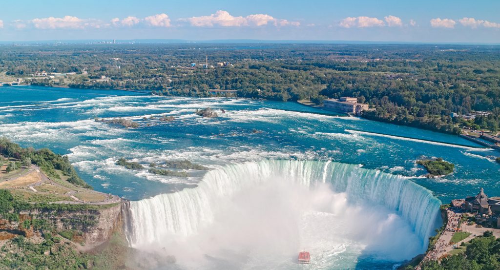 The thundering Horseshoe Falls crash dramatically into the Niagara River as a red tour boat approaches, surrounded by lush greenery.
