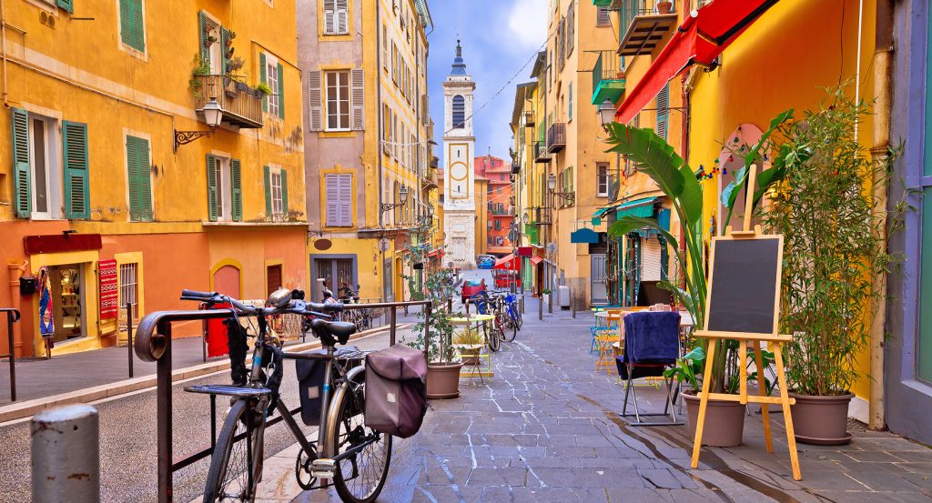 Charming city lane with outdoor seating, vibrant facades, and a distant bell tower under a blue sky.