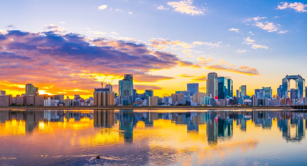 Colorful dawn over Osaka’s urban skyline with reflections of high-rise buildings and dramatic clouds in the water.
