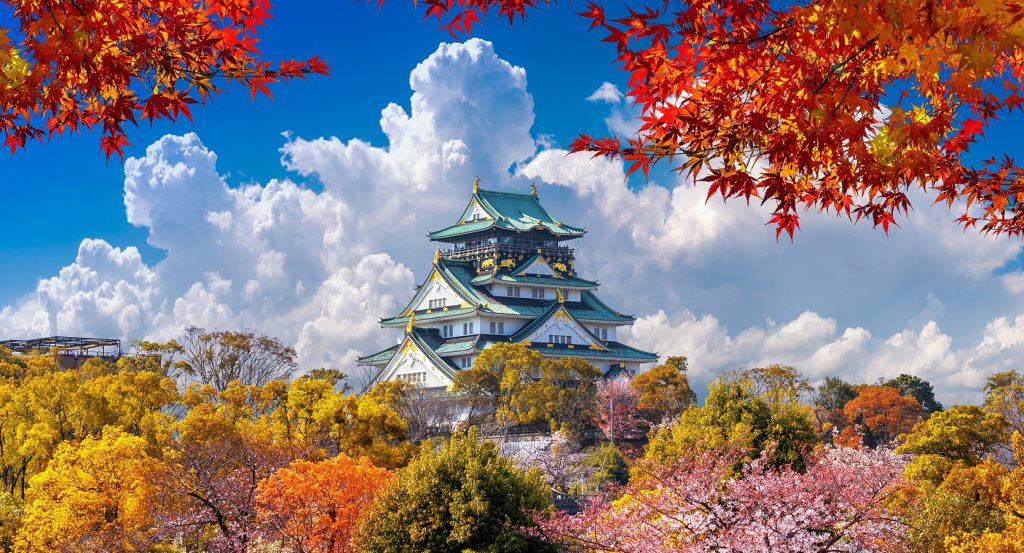 Osaka Castle surrounded by vibrant autumn foliage, cherry blossoms, and dramatic clouds under a brilliant blue sky in Japan.