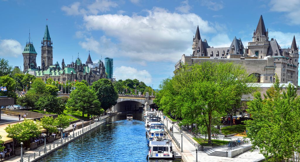 Boats line the calm waters of the Rideau Canal as Parliament Hill rises in the background beneath a blue sky.