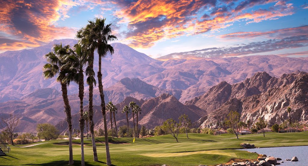 Palm trees and a golf course at sunset with rugged desert mountains in the backdrop, framed by fiery clouds.