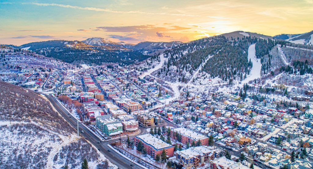 Aerial view of Park City covered in snow, with pastel-colored buildings lining the historic Main Street and surrounding ski slopes glowing at sunset.
