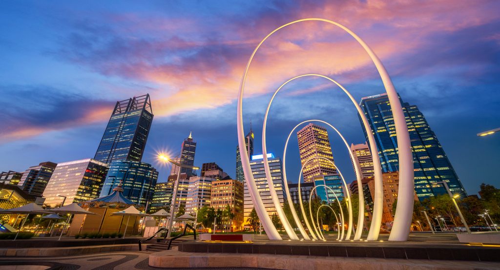 Perth city skyline at dusk featuring modern high-rises and the illuminated arches of Elizabeth Quay under a vibrant sunset sky.