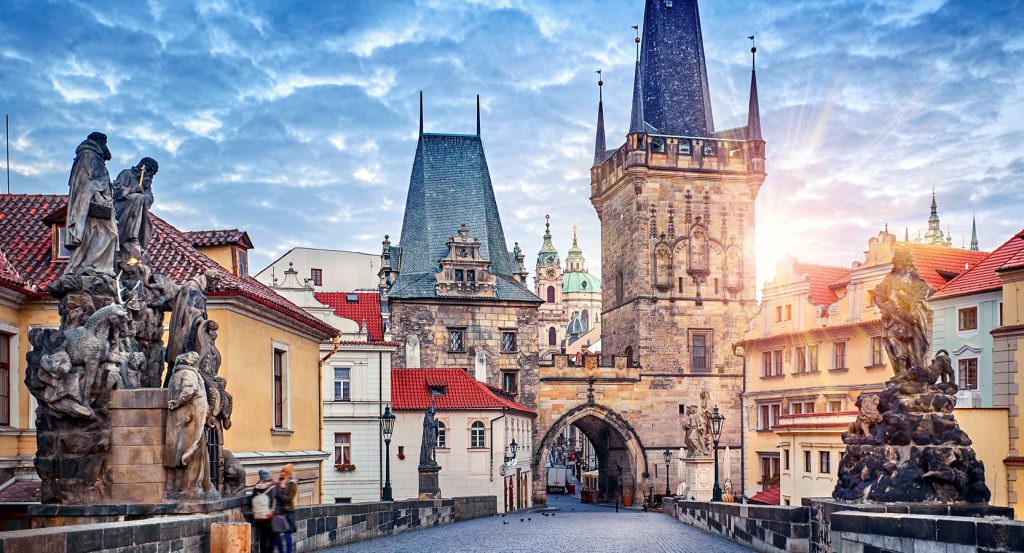 Sunlight peeking through the historic towers of Charles Bridge, framed by baroque statues and red-tiled rooftops in Prague.