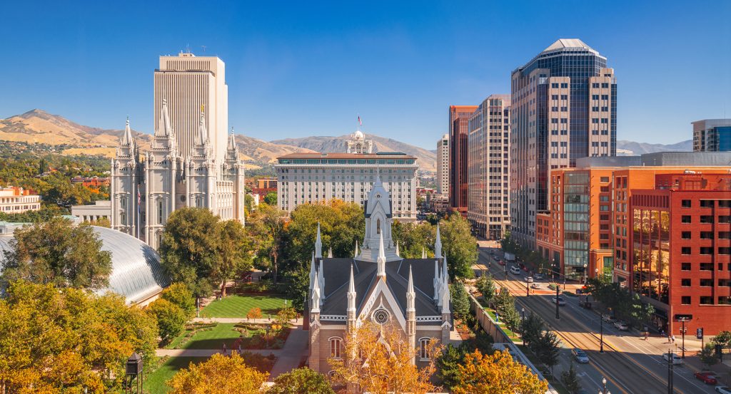 A sunny aerial view of Salt Lake City featuring the historic Salt Lake Temple and modern office towers against mountain peaks.