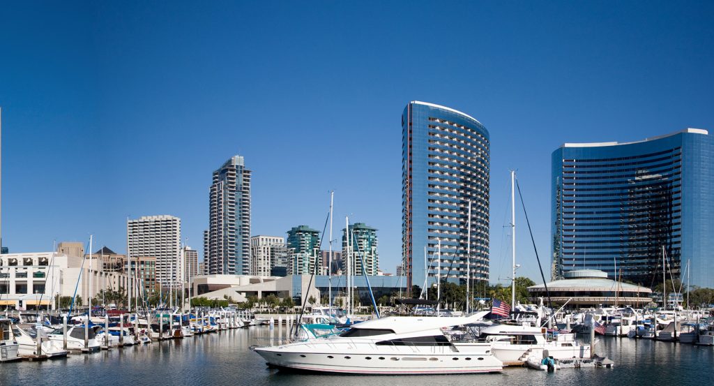 Luxury yachts docked at a marina with sleek, modern San Diego skyline rising behind under a clear blue sky.