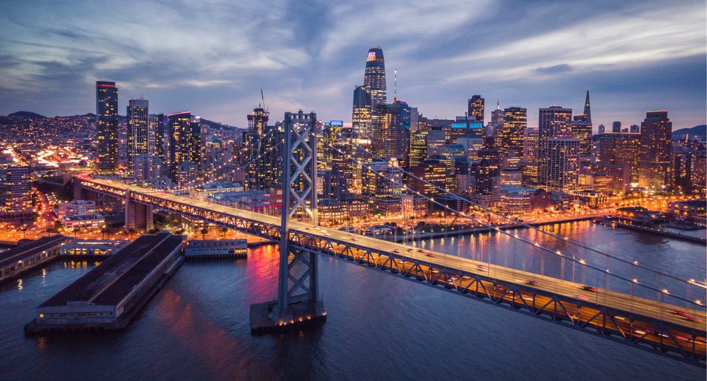San Francisco city lights and the Bay Bridge glowing at dusk, with skyscrapers stretching into the skyline.