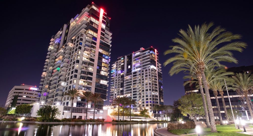 Glowing high-rise hotels reflect off a tranquil night-lit waterway, framed by tall palms in Santa Ana’s sleek urban core.