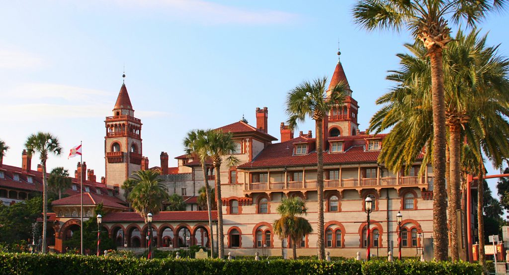 A majestic red-roofed building with towers and palm-lined gardens, basking in the soft evening light of St. Augustine.
