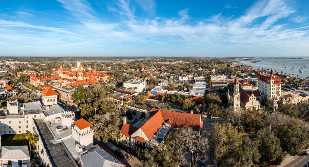 Aerial view of St. Augustine’s historic red-roofed architecture with coastal waters glinting in the distance.