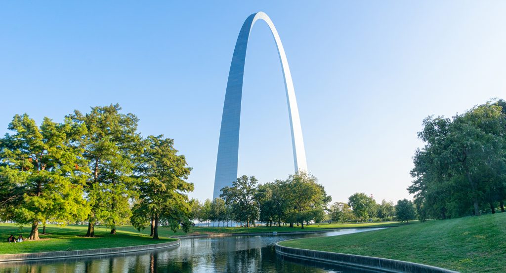 St. Louis’ iconic Gateway Arch surrounded by trees and tranquil water under a blue sky.