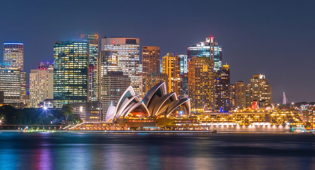 Vibrant waterfront view of Sydney Harbour with city lights reflecting on the water and the Opera House in the foreground.