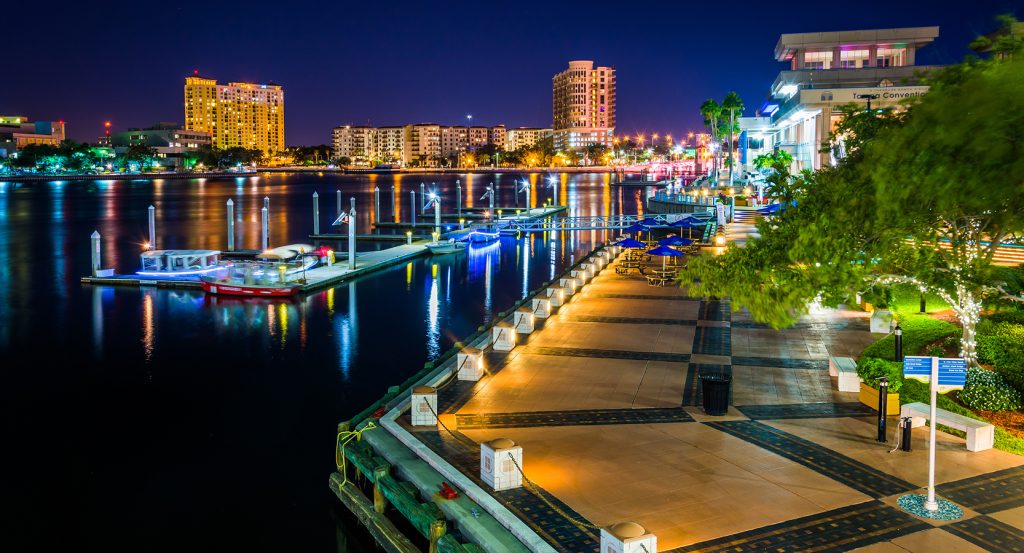 Nighttime view of downtown Tampa’s Riverwalk, featuring a well-lit marina, modern buildings, and festive trees.