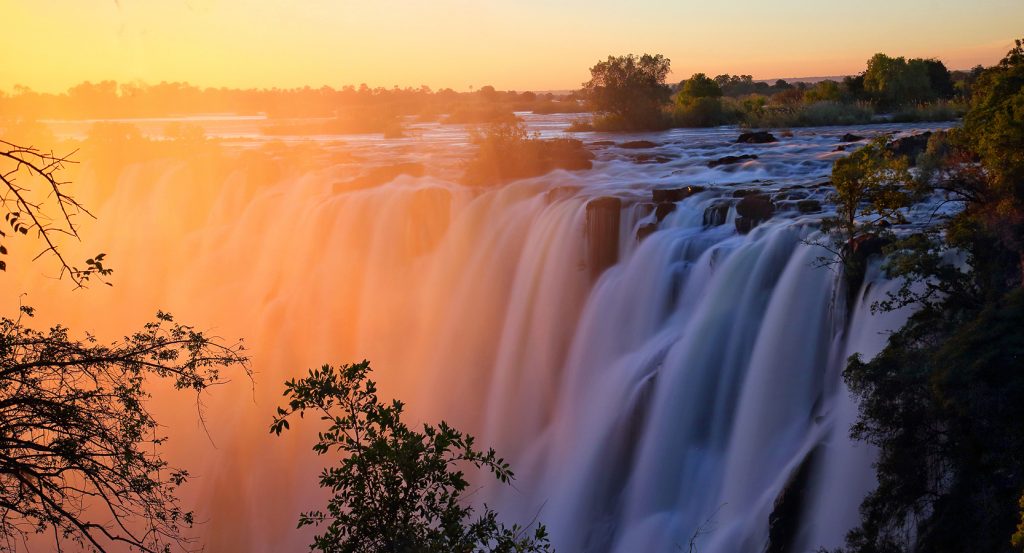Golden light illuminates the roaring waters of Victoria Falls at dawn, surrounded by trees and cliffs.