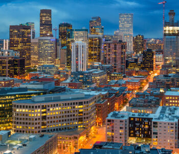 Downtown Denver skyline glowing at dusk with illuminated high-rise buildings and city lights stretching across the Mile High City.