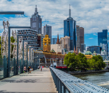 Pedestrians crossing a modern riverside walkway with Melbourne’s central skyline rising behind them.