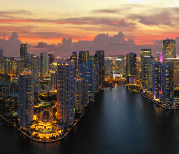 Panoramic aerial view of the Miami skyline at sunset, with glowing skyscrapers rising above Biscayne Bay.