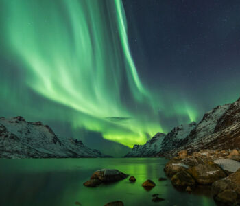 The Northern Lights ripple in green waves over a snow-covered fjord flanked by jagged mountains near Reykjavík, Iceland.