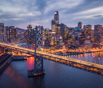 Aerial evening view of San Francisco’s skyline with the Bay Bridge illuminated against a twilight sky.