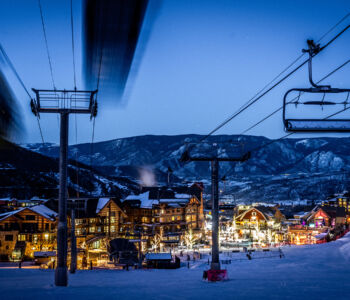 Ski lift gliding over Vail Village at dusk, with glowing chalet lights against snow-covered mountains.