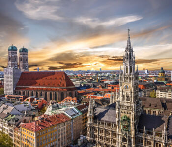 Aerial view of Munich skyline featuring Frauenkirche towers and Neues Rathaus at sunset.