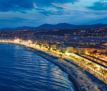 A sweeping view of the Promenade des Anglais, lined with pastel buildings and palm trees beside the azure Mediterranean.
