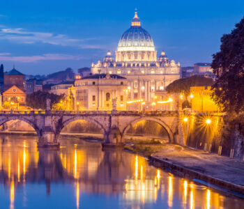 St. Peter’s Basilica illuminated at dusk above the Tiber River, with Ponte Sant’Angelo reflected in the water.