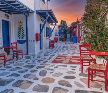 Colorful cobblestone street in Ano Mera, Mykonos with red chairs and blue doors at sunset.