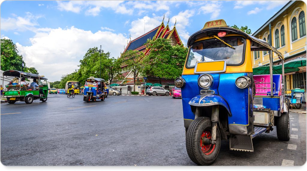 Blue tuk tuk taxi near Wat Pho in Bangkok, parked in front of a temple and bustling street corner.