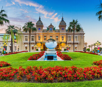 Casino de Monte-Carlo with the Sky Mirror fountain, manicured lawn, red flower beds, and palm trees at blue hour in Monaco
