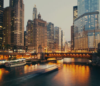 Chicago Riverwalk at dusk with illuminated skyscrapers, a steel bridge glowing over the river, and boats moving along the water.