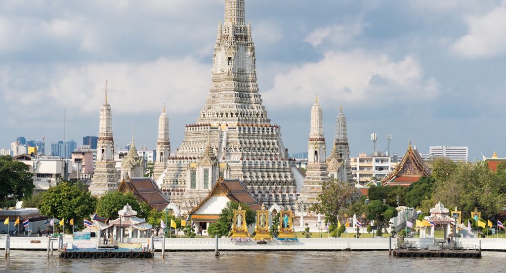 A riverside view of Wat Arun’s intricate architecture framed by fluttering flags and lush greenery in Bangkok, Thailand.