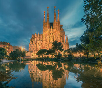 The Sagrada Família at dusk reflected in a calm pond, with dramatic blue skies and warm golden lighting.