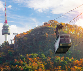 Cable car ascending Namsan Mountain with N Seoul Tower rising behind autumn-colored trees in Seoul.