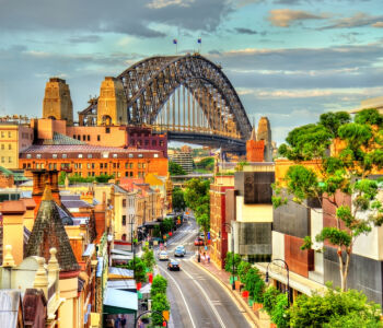 View of The Rocks district in Sydney with heritage buildings in the foreground and the Sydney Harbour Bridge rising dramatically behind them under a soft, golden sky.