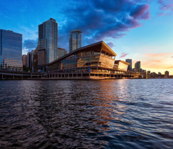 The Vancouver Convention Centre lit by sunset, with downtown skyscrapers rising behind it over the waterfront.