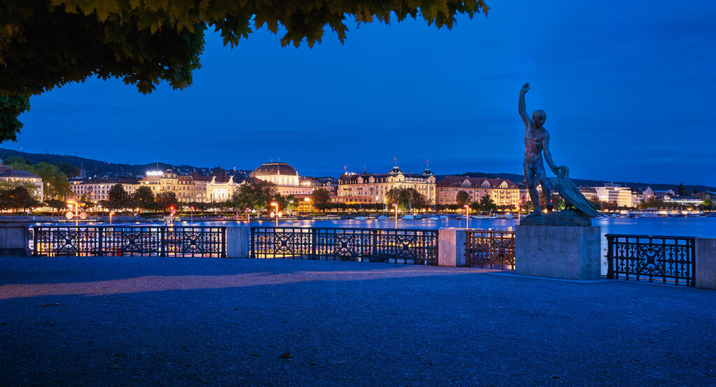 Nighttime view of Bürkliplatz with statue and lights reflecting on Lake Zurich