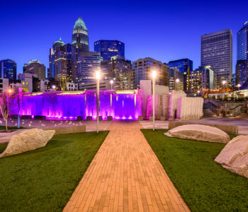 Bearden Park in Charlotte illuminated at blue hour with glowing fountains and the uptown skyline rising behind it.