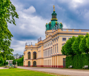 Charlottenburg Palace in Berlin, Germany, with its ornate Baroque architecture, green copper dome, and surrounding gardens under a dramatic cloudy sky.