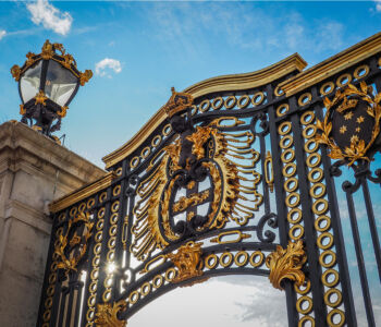 Ornate black and gold gates outside Buckingham Palace with intricate royal crests against a bright blue sky.