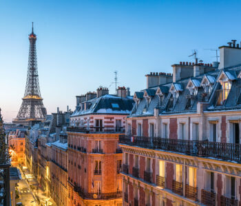 Eiffel Tower rising above Parisian rooftops at dusk, viewed from a classic Haussmann-style street.